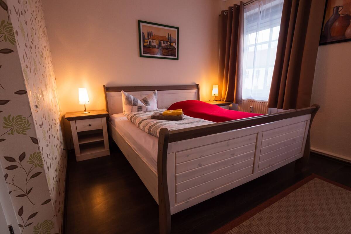 A neatly arranged bedroom with a wooden bed, two bedside tables, a nightstand, a framed picture on the wall, and a window with curtains.