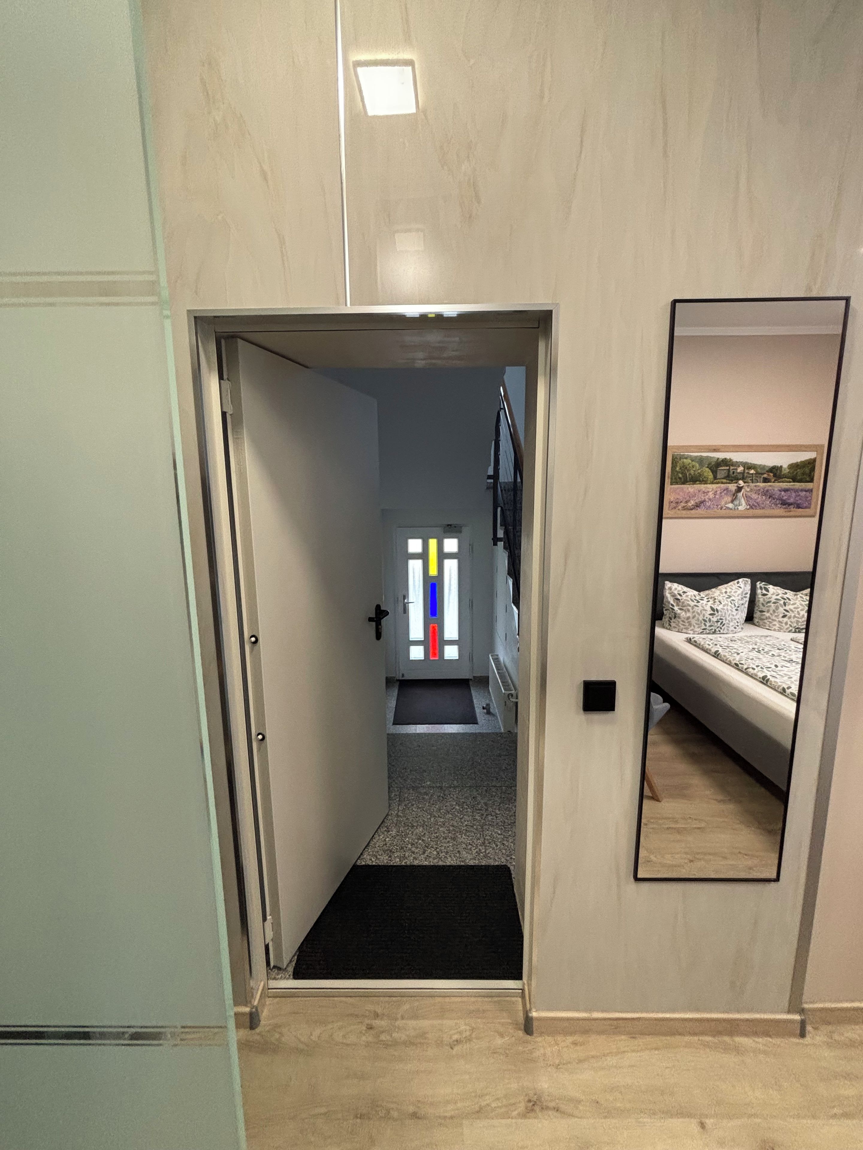 A narrow hallway with a door leading to a room with a bed and a mirror reflecting a room with a bed.