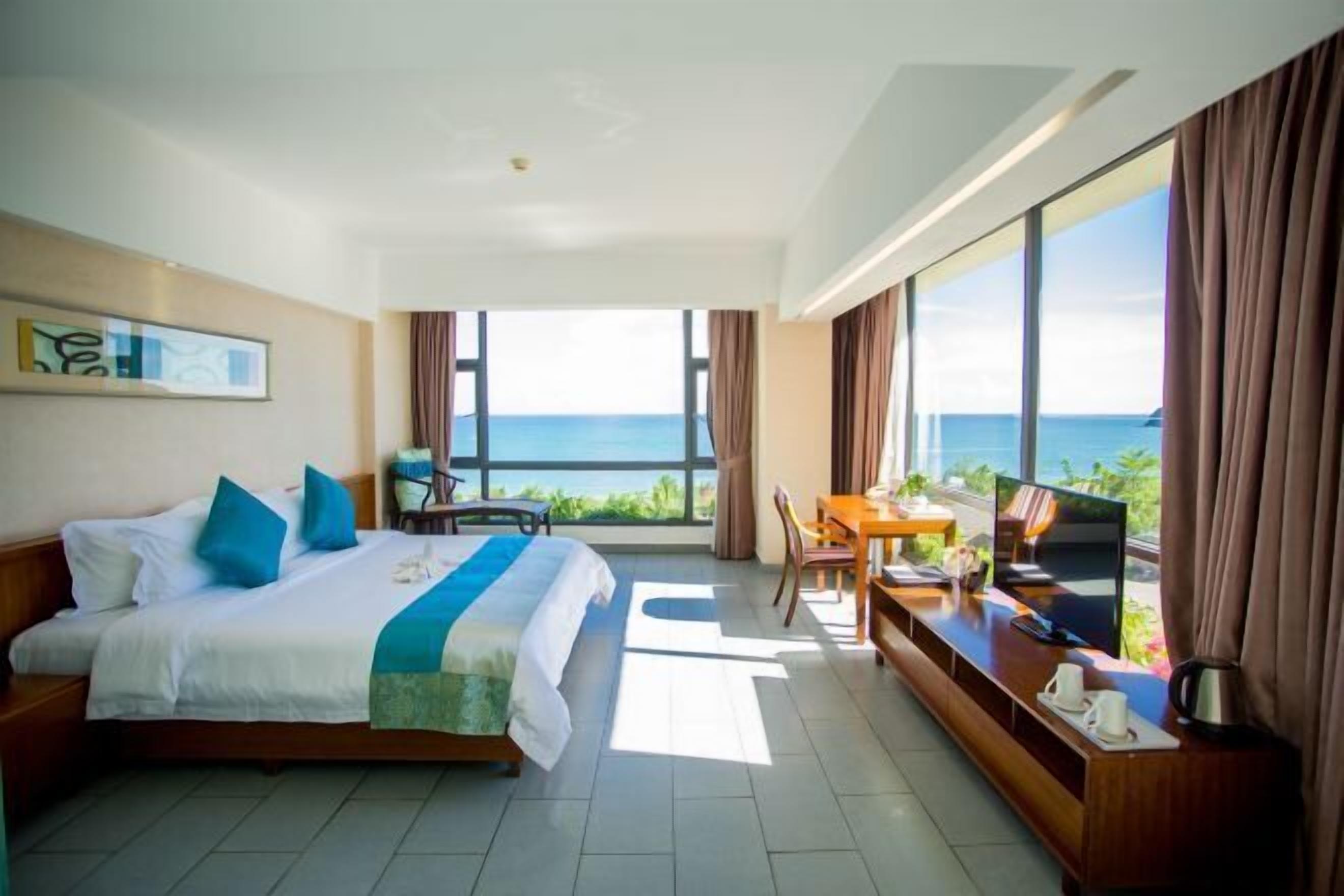 A hotel room with a bed, a desk with a TV, and a view of the ocean.