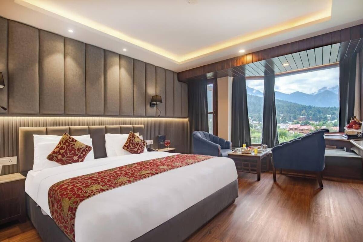 A hotel room with a large bed, a desk with a chair, a view of the mountains, and a cityscape.