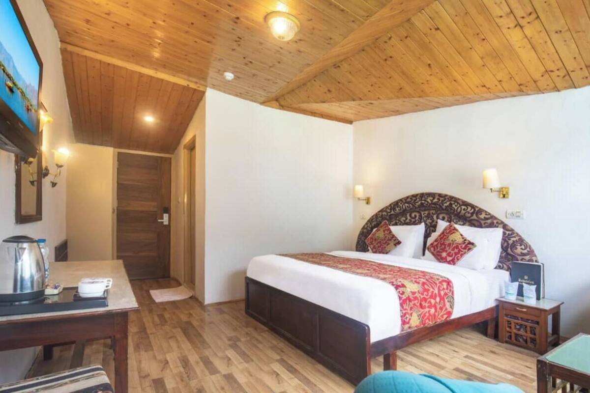 A bedroom with a wooden ceiling, a bed with patterned headboard, bedside tables, and a television on the wall.