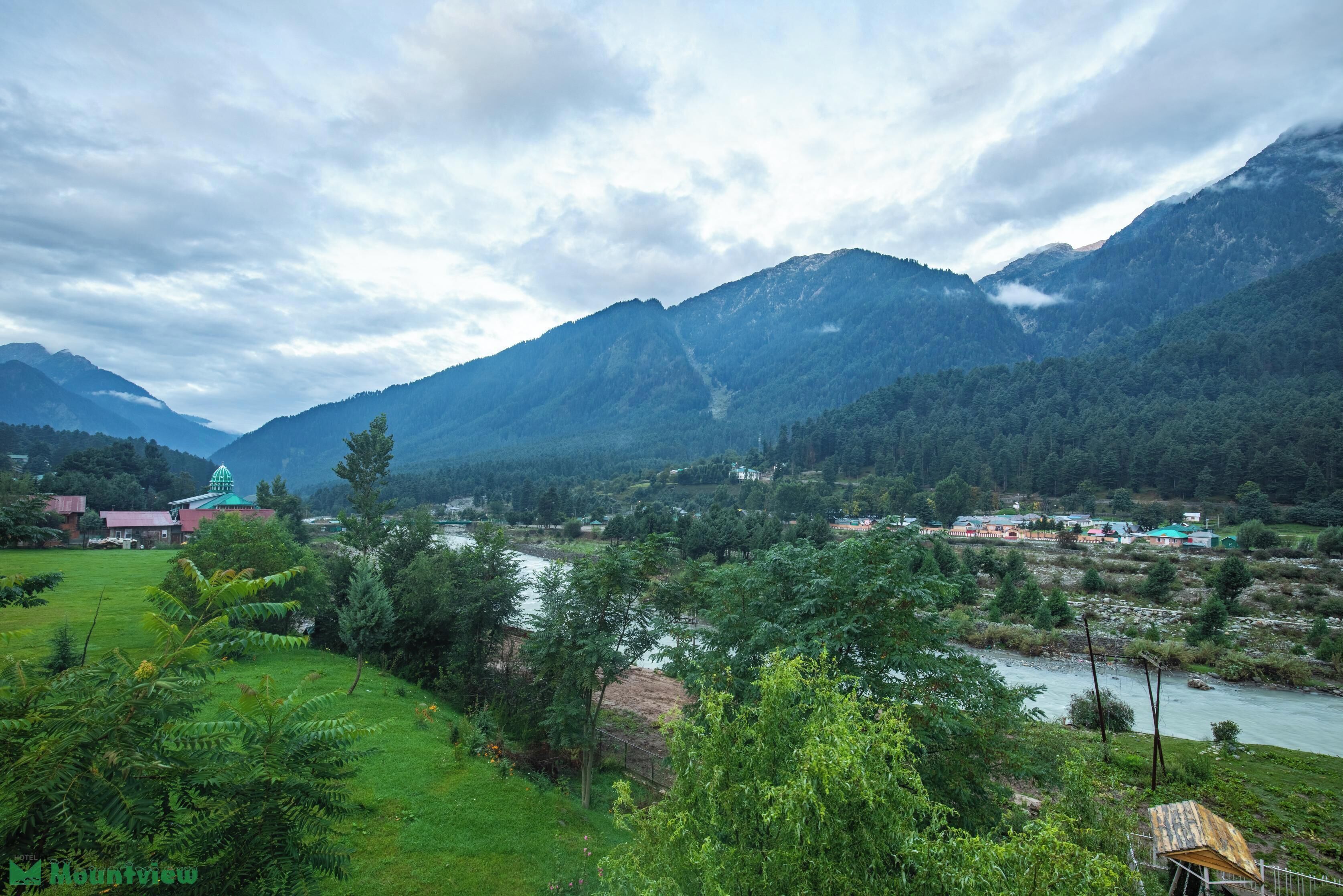 A mountainous landscape with a river, greenery, and a building with a green roof.