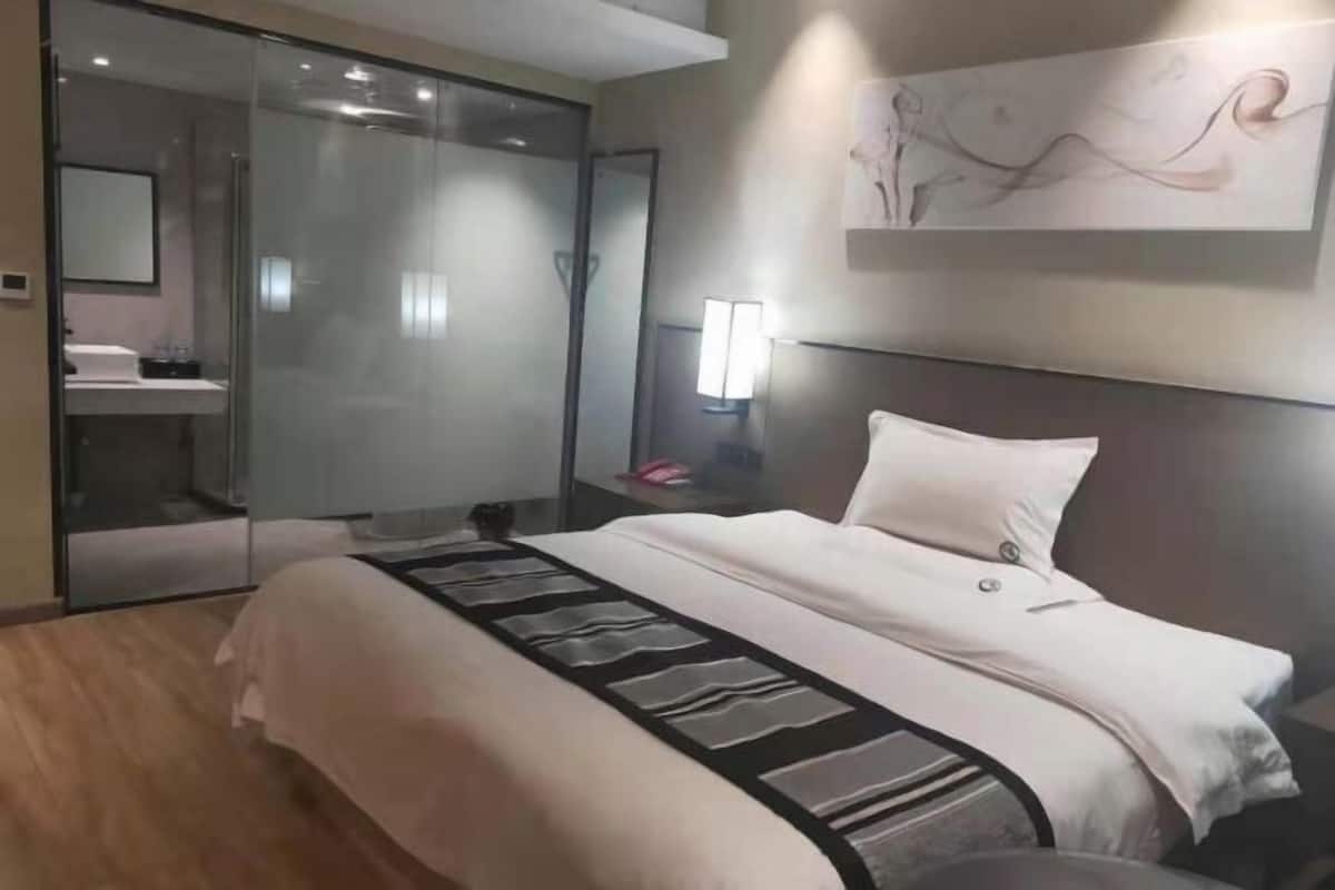 A modern hotel room with a large bed, a glass shower enclosure, a bedside table with a lamp, and a wall-mounted artwork.
