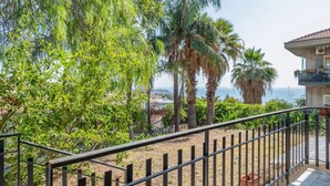 Un balcón con barandilla metálica, que da a un jardín con varios árboles y vistas al mar.