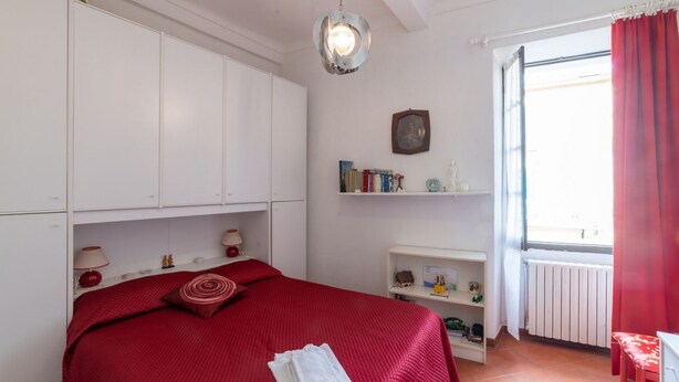 A bedroom with a bed, a nightstand, a lamp, a shelf with books, a window with curtains, and a radiator.