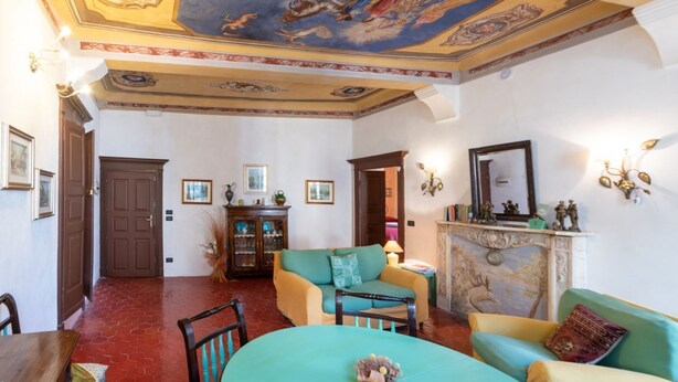 A room with a dining table, chairs, a sofa, a fireplace, and a ceiling with a fresco.