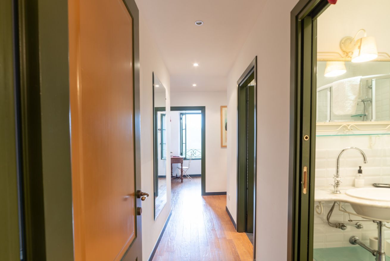 Un couloir avec un plancher en bois, une porte donnant sur une salle de bain équipée d’un lavabo et d’un miroir, et une fenêtre à l’arrière-plan.