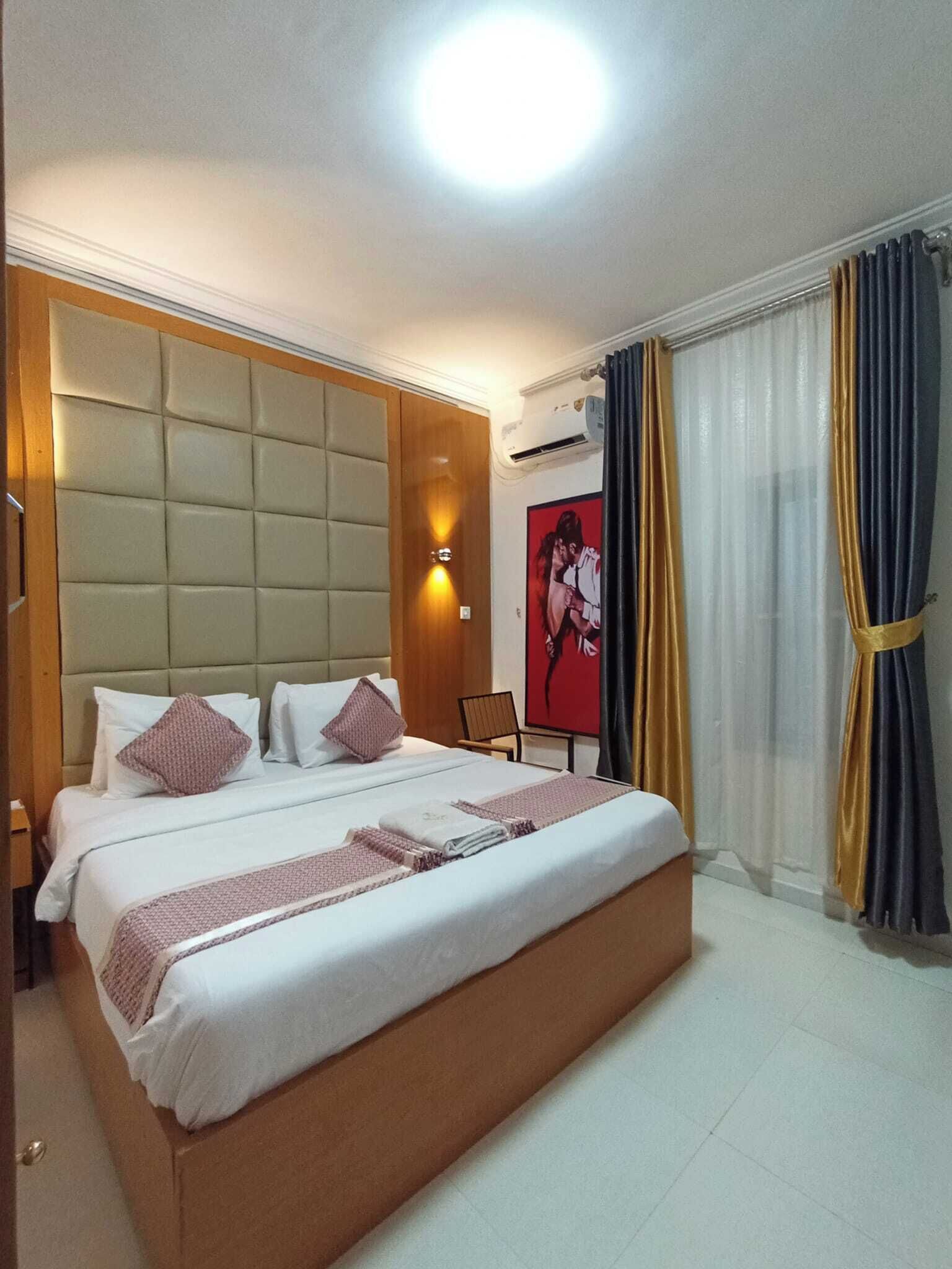 Une chambre d’hôtel avec un grand lit, une tête de lit, une table de chevet, une chaise, un climatiseur fixé au mur et une fenêtre avec des rideaux.