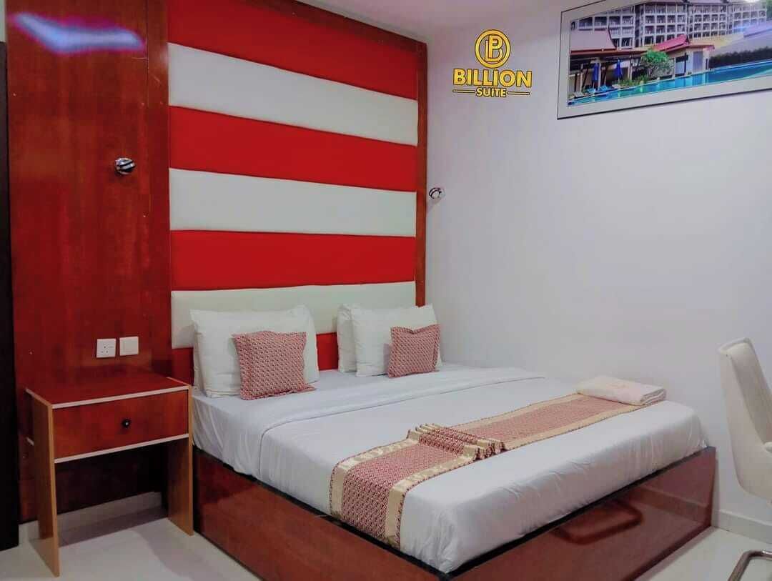 A hotel room with a large bed, a wooden nightstand, a white chair, and a red and white striped headboard.