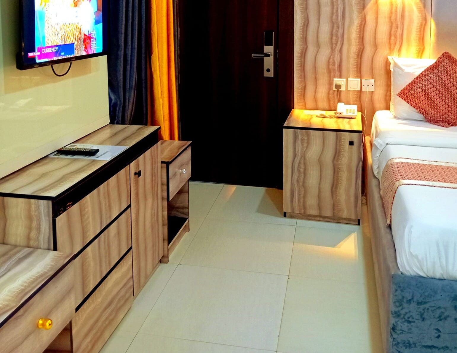 Una camera d'albergo con una scrivania in legno, un letto con testiera imbottita blu, un comodino con una lampada e una TV a parete.