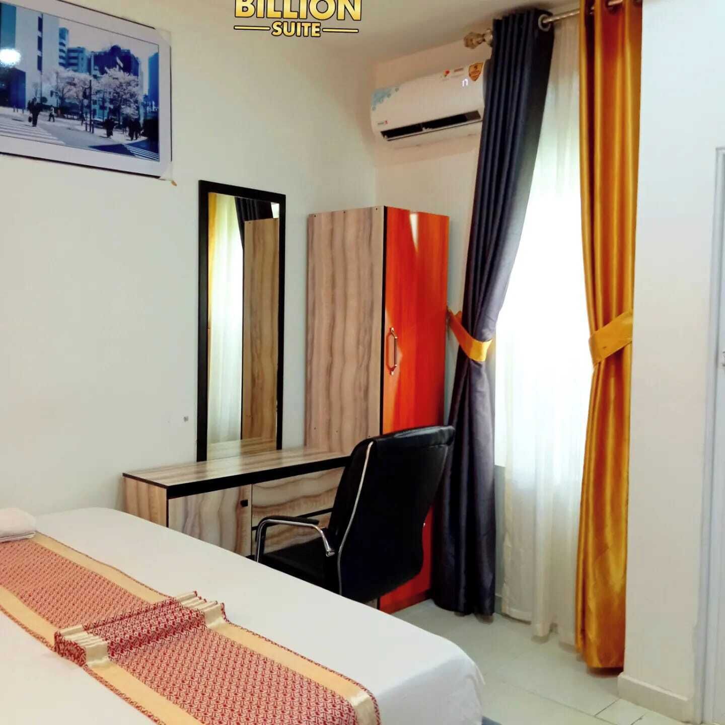 A hotel room with a bed, a desk, a chair, a wardrobe, a mirror, and an air conditioning unit.