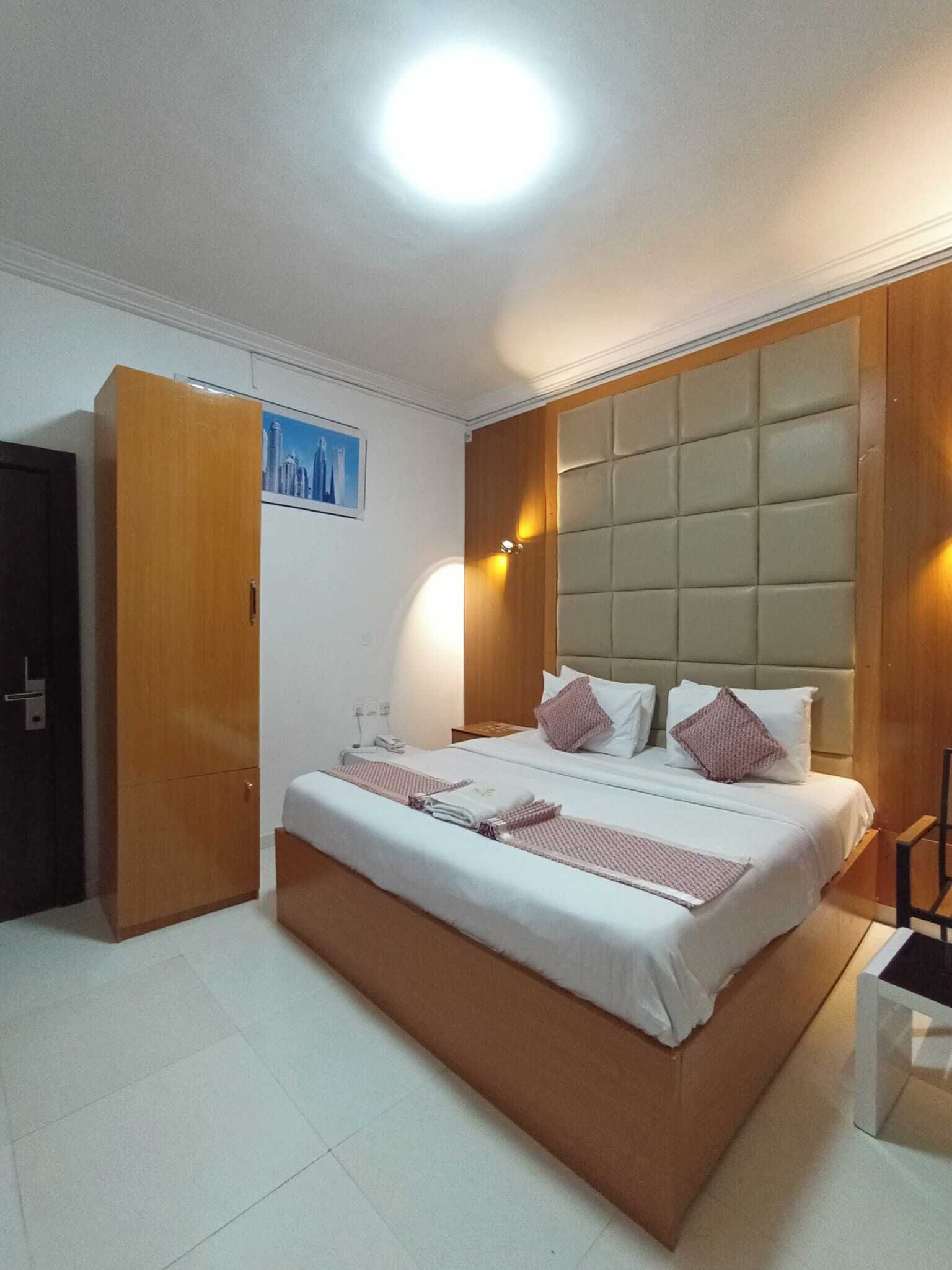 A hotel room with a large bed, a wooden wardrobe, a cityscape picture on the wall, and a padded headboard.