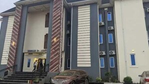 Un edificio a più piani con un ingresso centrale, condizionatori d'aria e un'area di parcheggio con auto.
