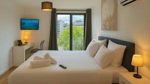Ein Hotelzimmer mit einem großen Bett, einem Schreibtisch mit Lampe, einem Fernseher und Blick auf Gebäude und Bäume durch das Fenster.