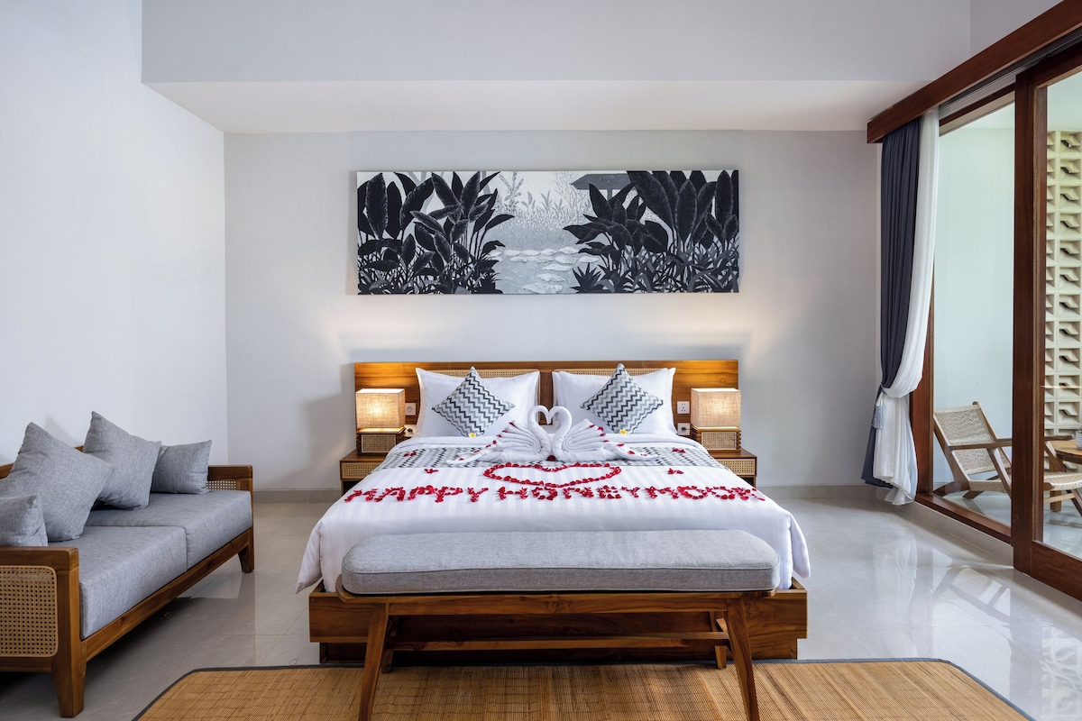 A hotel room with a large bed, a sofa, a wall-mounted artwork, and a view through a glass door.