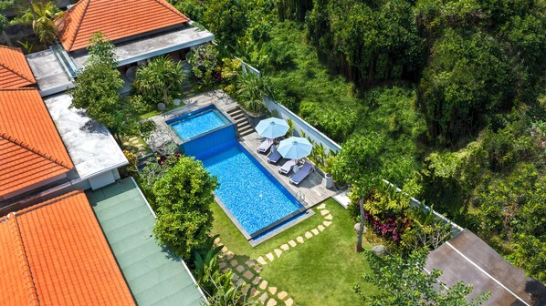 Vista aérea de una propiedad con piscina rectangular, rodeada de exuberante vegetación y una terraza de madera con sillones y sombrillas.