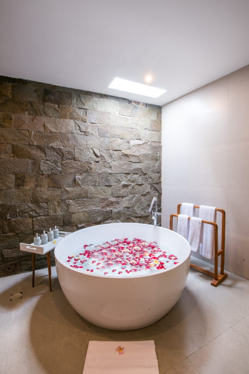 A modern bathroom with a stone wall, a freestanding bathtub filled with rose petals, a small table with toiletries, and a towel rack.