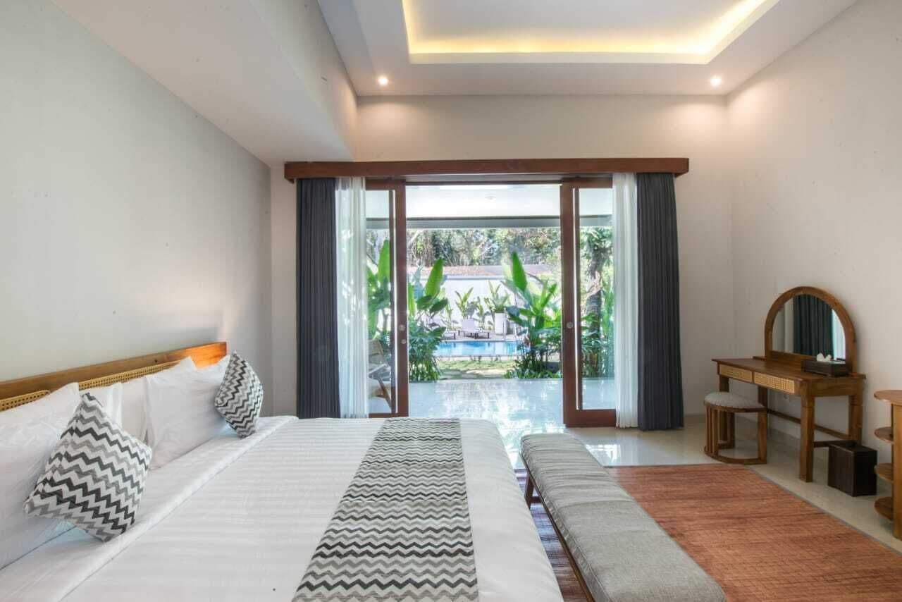 Une chambre avec un grand lit, un banc, une commode en bois et une vue sur une piscine et la verdure environnante.