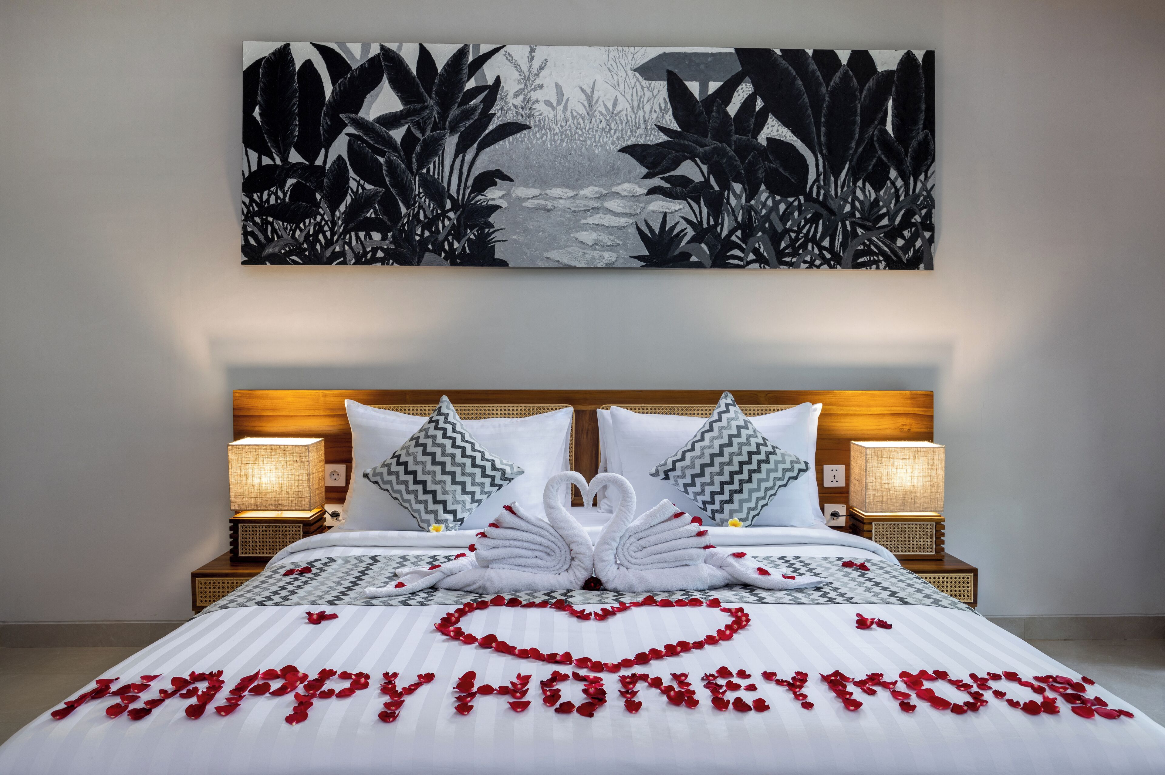 A hotel room with a bed adorned with a heart-shaped arrangement of rose petals spelling "HAPPY HOME" and two swan-shaped towels.