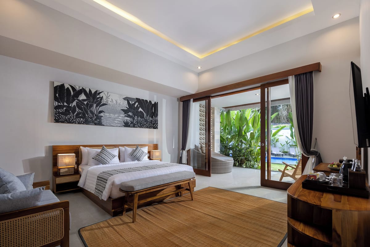 A modern hotel room with a large bed, a sitting area, a dining table, and a view of a pool and greenery.