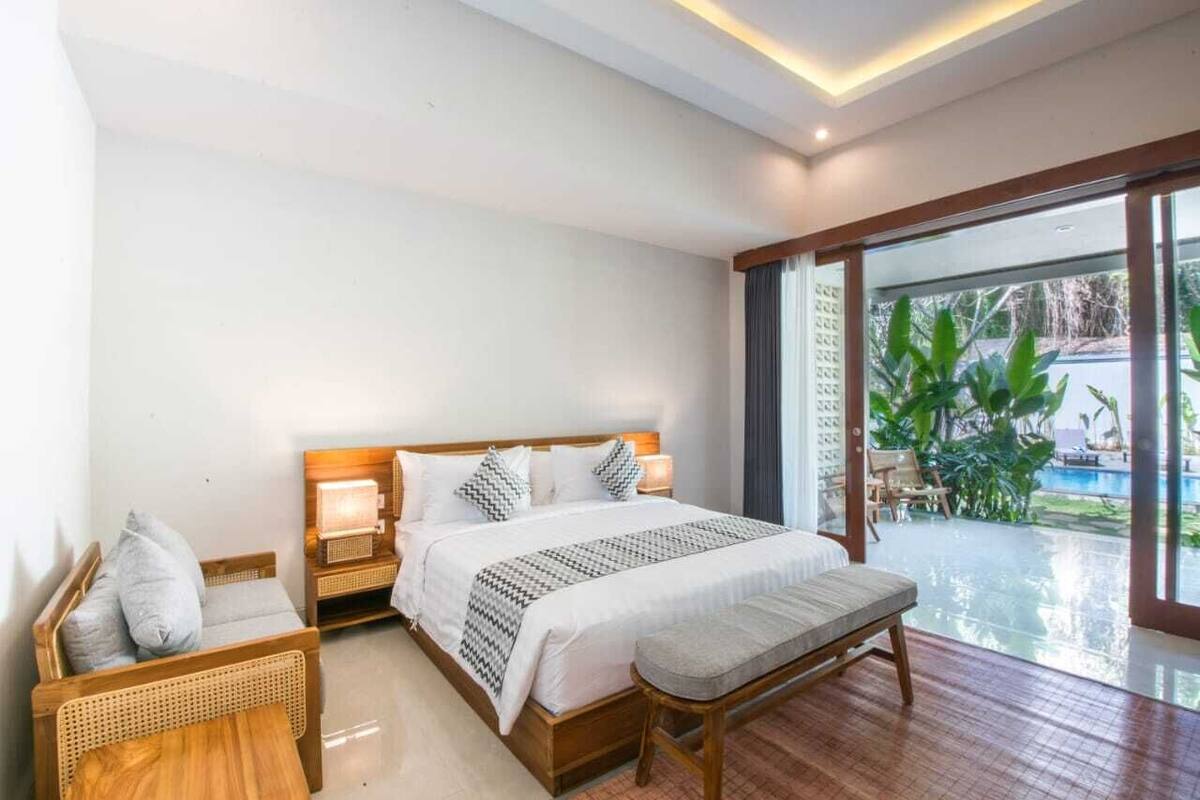 A bedroom with a large bed, bedside tables, a bench, and a view of a pool through a glass door.