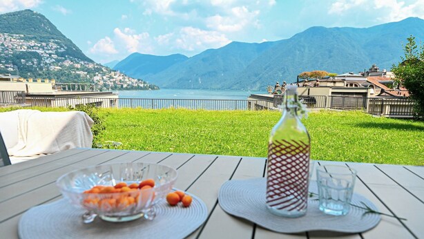 A table with a bottle, a glass, and a bowl of fruit, overlooking a scenic view of a lake and mountains. - Luxury Central Suite Garden in Lugano (Lugano)