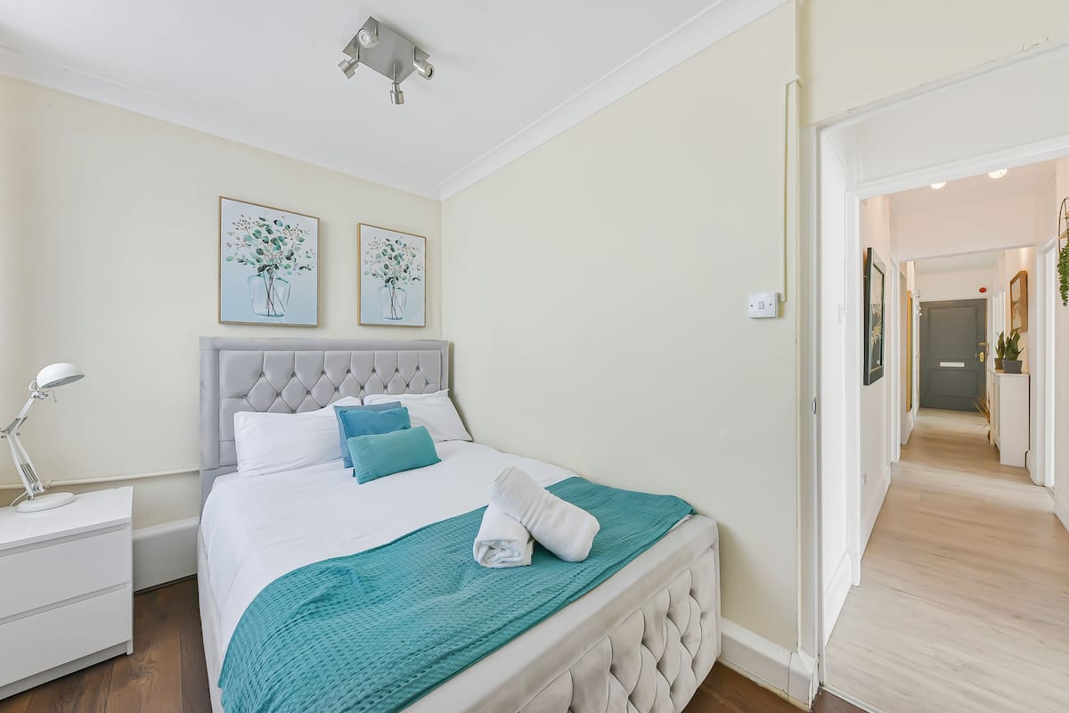 A neatly made bed with a white headboard, a teal throw blanket, and a folded towel. There is a white bedside table with a lamp and a framed picture on the wall above the bed.