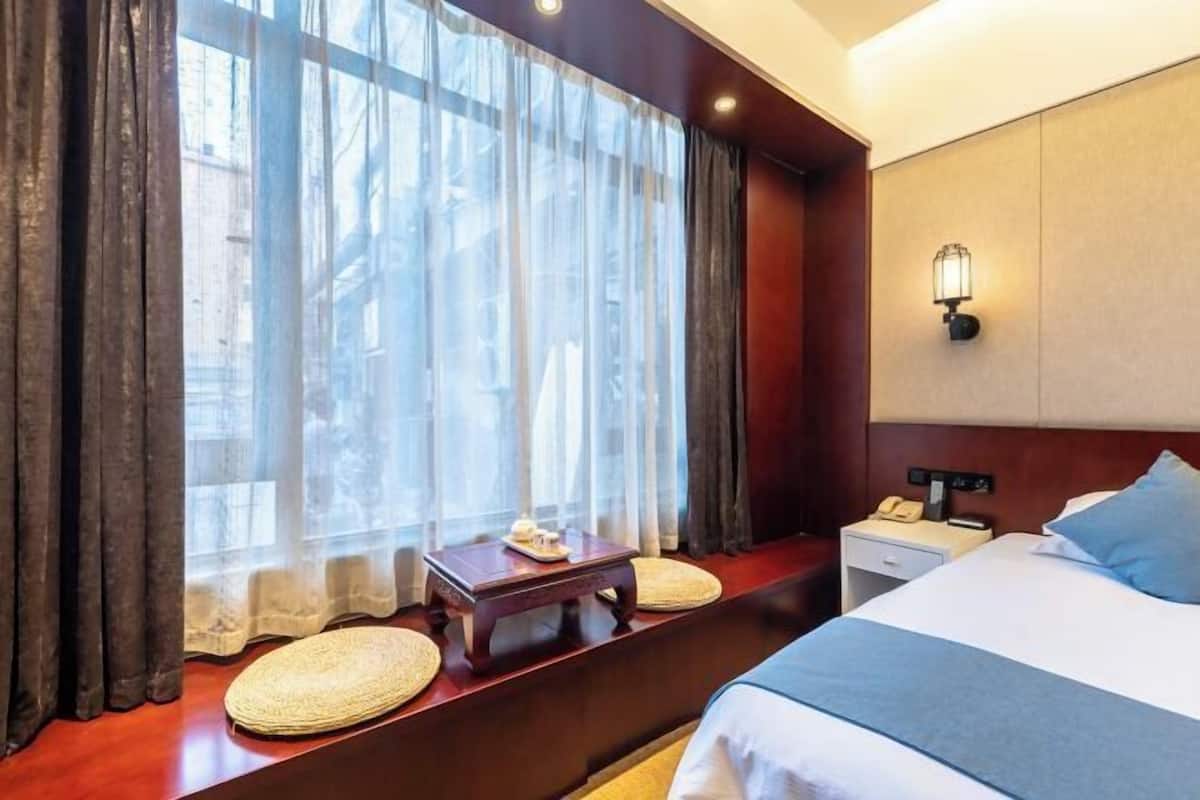 Habitación de hotel con cama, mesita de noche, lámpara, ventana con cortinas y vistas a edificios.