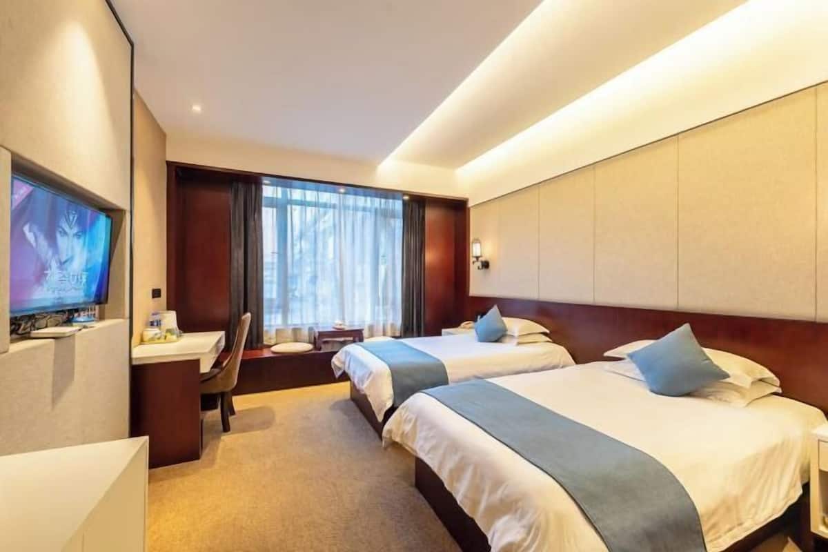 Habitación de hotel con dos camas, un escritorio, un televisor y un ventanal grande con cortinas.