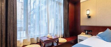A hotel room with a large bed, a bedside table with a lamp, a small table with a plate of food, and a window with sheer curtains.