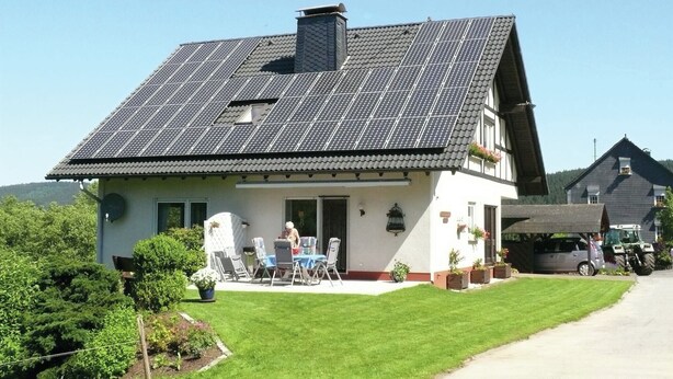 Una casa con techo inclinado, paneles solares, un porche cubierto con sillas y césped.