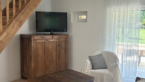 Una sala de estar con una escalera de madera, un televisor sobre un mueble bajo, un sofá blanco y vistas a través de una ventana.