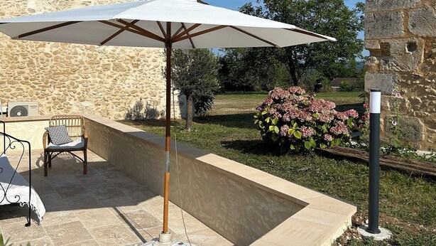 Un patio con muro de piedra, una sombrilla blanca, una silla de mimbre y una mesita.