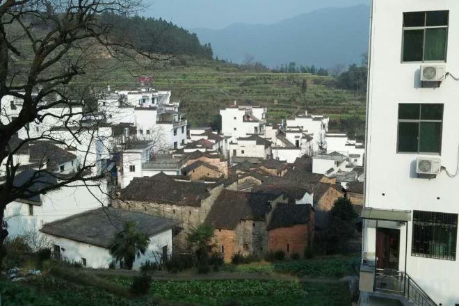 A village with traditional white buildings, a modern building with air conditioning units, and a hilly landscape in the background.