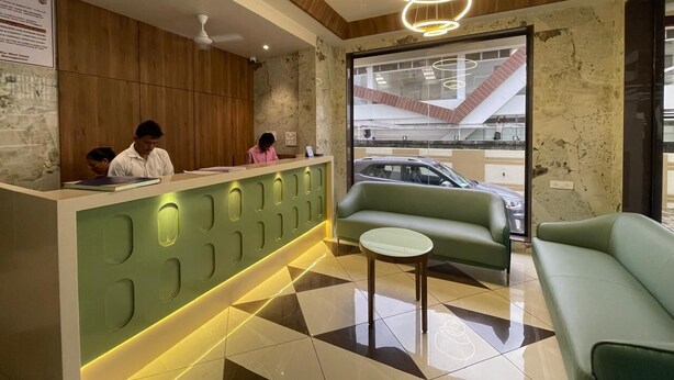 A reception area with a green counter, two receptionists, a green sofa, a small table, and a view of the outside through a large window. - Hotel The West Coast (Vasai)