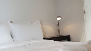 Un letto rifatto con lenzuola bianche, una lampada da comodino e un comodino scuro in una stanza semplice e pulita.