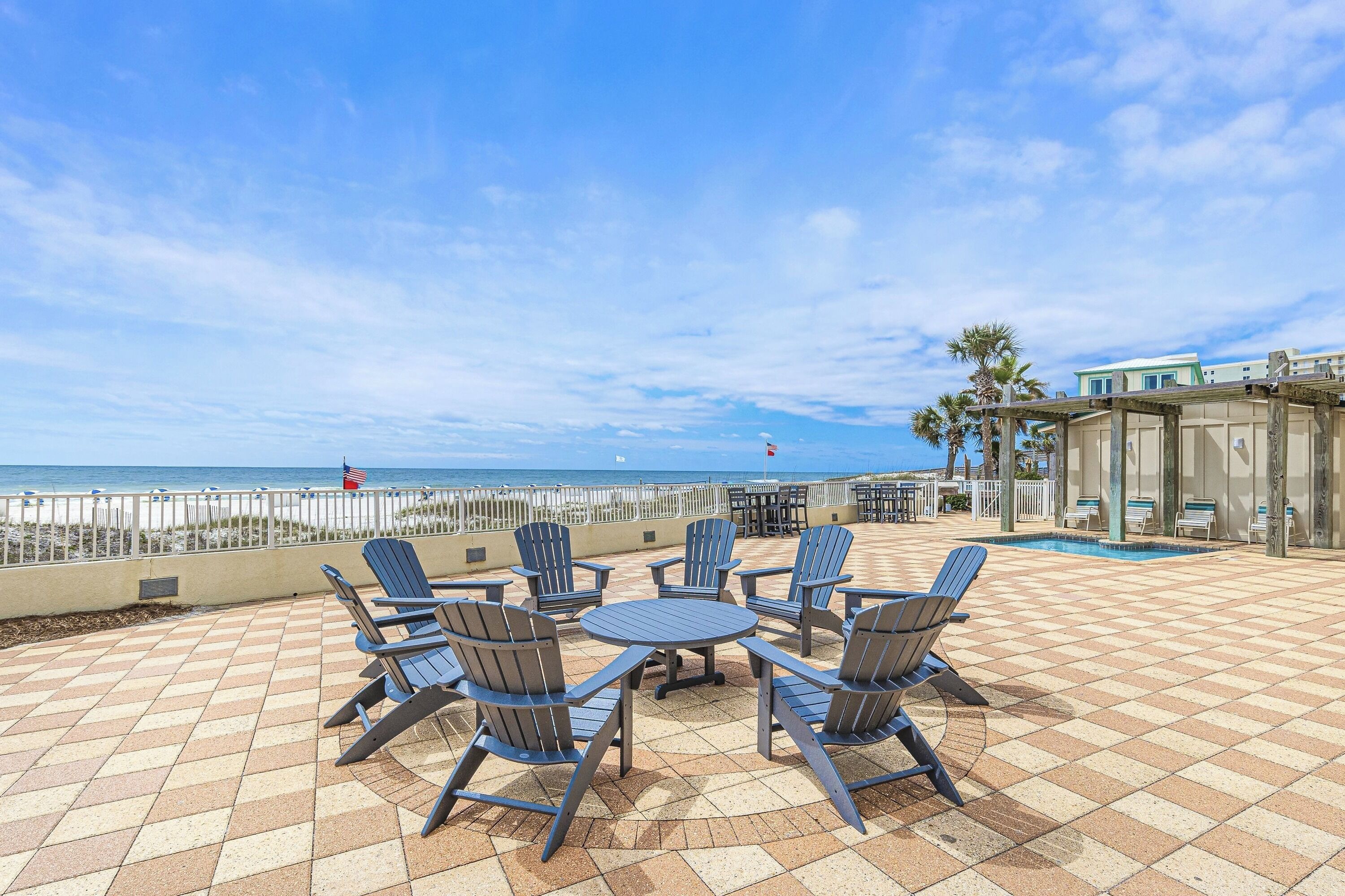 A patio with Adirondack chairs and a pool table, overlooking a beach with a fence and palm trees.
