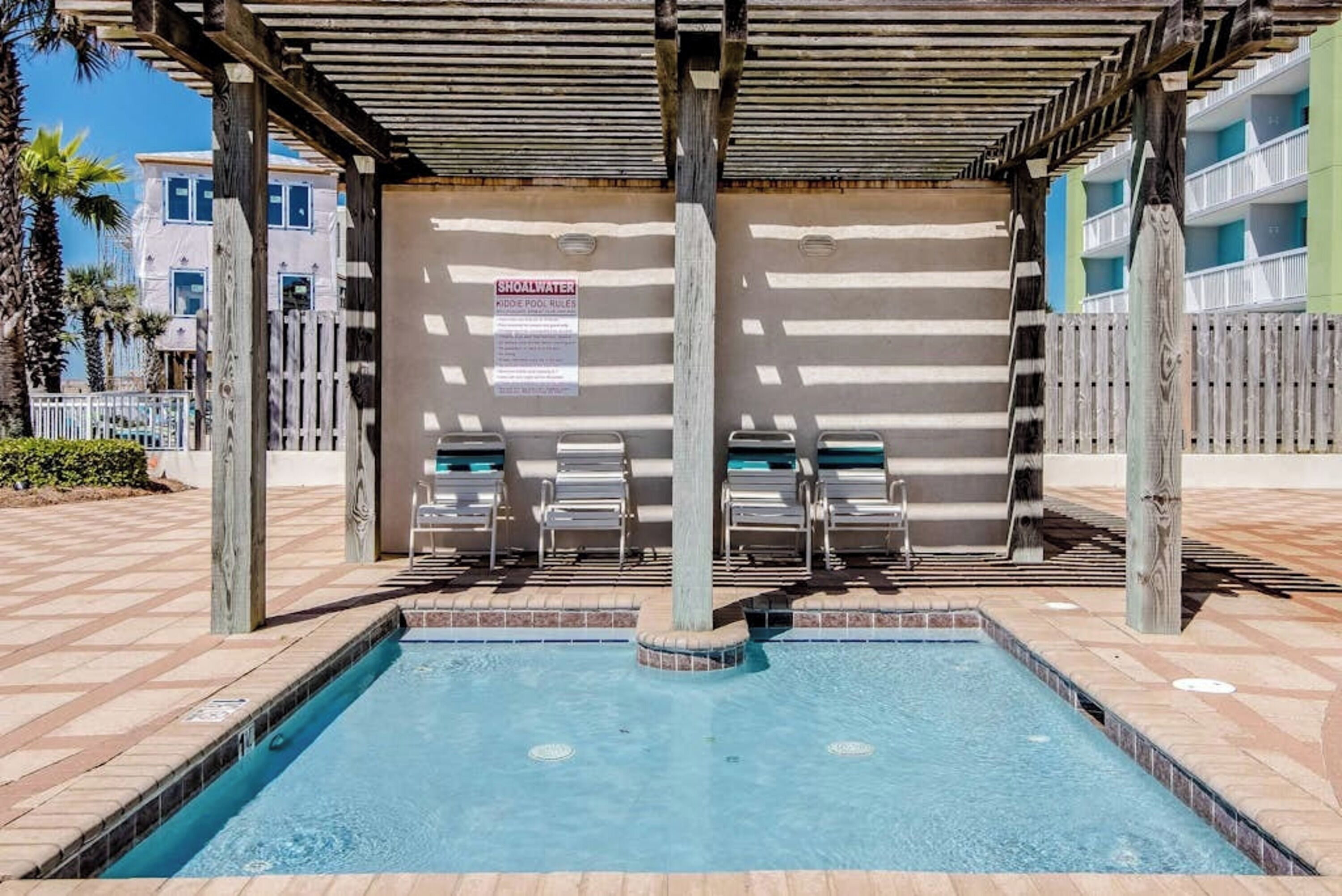 A pool area with a covered seating area, a sign indicating kiddie pool rules, and a wooden fence.