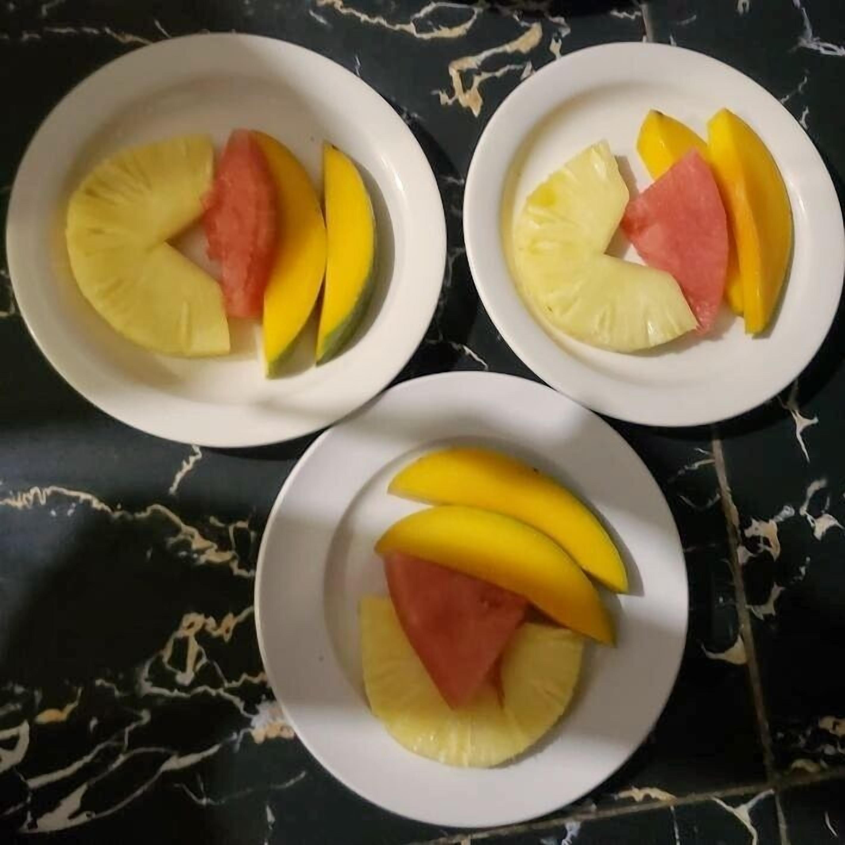 Three white plates with slices of pineapple, mango, and watermelon arranged on a dark marble surface.