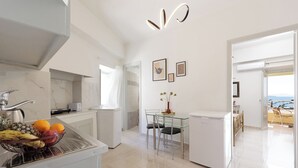 Une cuisine moderne dotée d’un îlot central, d’un coin repas avec une table et des chaises en verre, et offrant une vue sur la mer grâce à une porte ouverte.