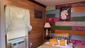 Un dormitorio con cabecera de madera, una cama con ropa de cama estampada, una mesita de noche con lámpara y una pared con un mural colorido.