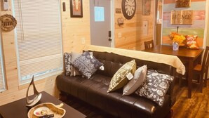Una sala de estar acogedora con un sofá de cuero negro, una mesa de comedor con sillas, y una pared de madera con un reloj y fotografías enmarcadas.