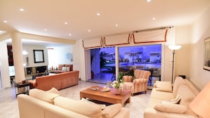 A spacious living room with a large sofa, a coffee table, and a TV. There is a sliding glass door leading to a balcony with a view of houses.