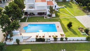 A large swimming pool with lounge chairs, surrounded by a well-maintained lawn and a white building with a tiled roof.