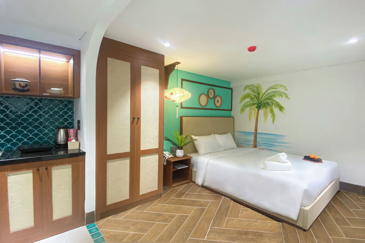 A hotel room with a bed, a nightstand, a lamp, a wall with a palm tree mural, and a kitchen area with cabinets and a tiled backsplash.