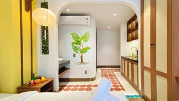 A room with a bed, a bedside table with fruits, a wall-mounted air conditioner, and a kitchen area with cabinets and a sink.