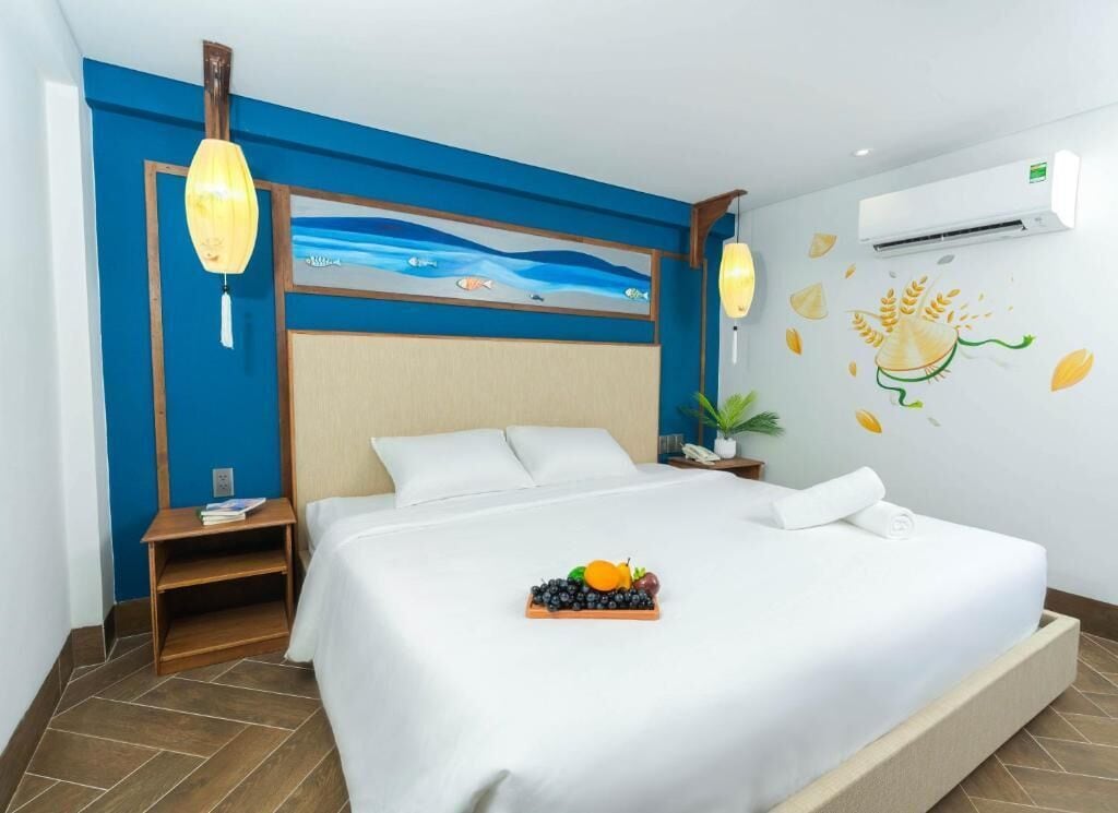 A hotel room with a bed, a nightstand, a wall-mounted air conditioner, and a wall decoration of fruits and a sun.