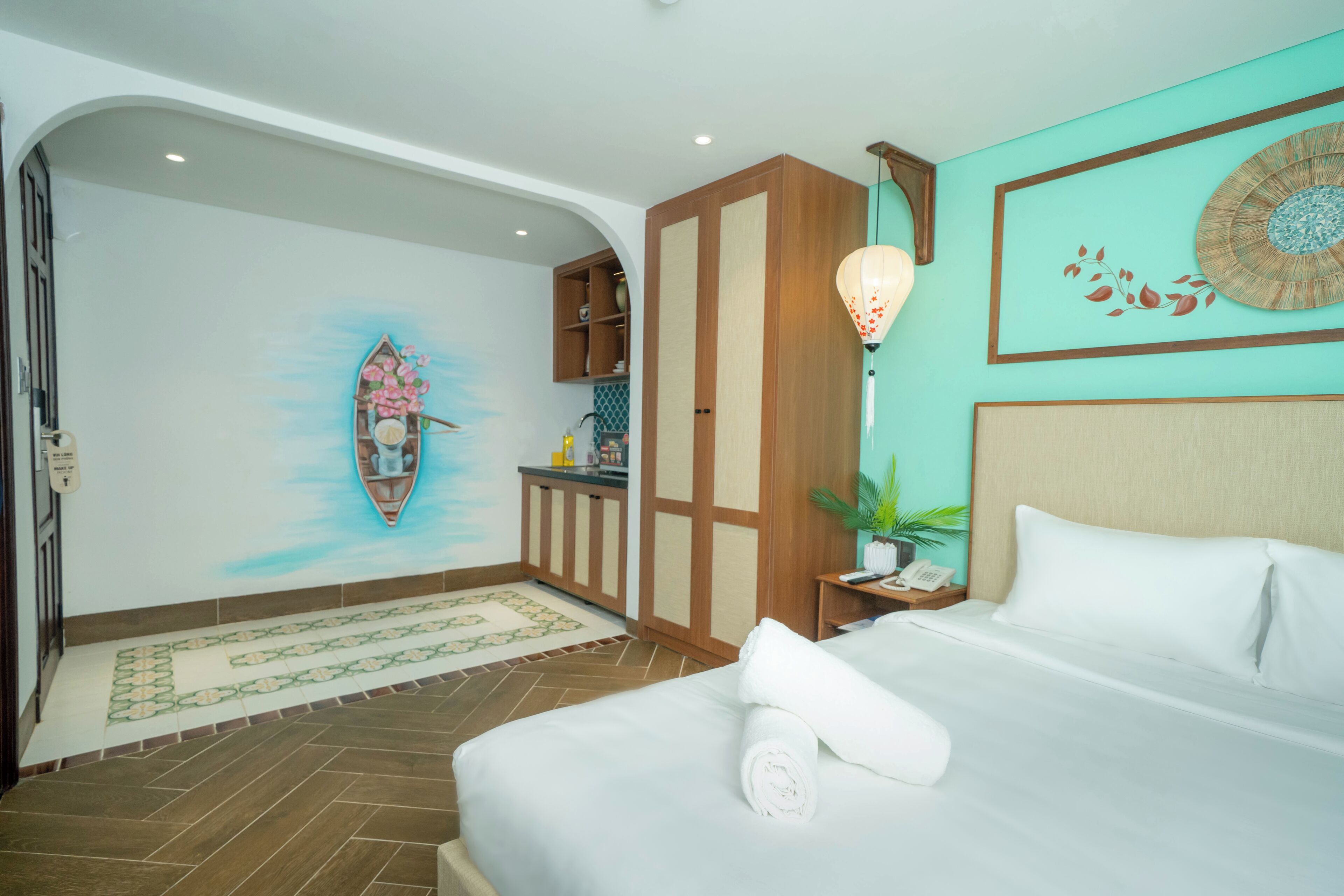 A hotel room with a bed, a nightstand, a wall-mounted lamp, a small table with a plant, and a decorative wall piece.