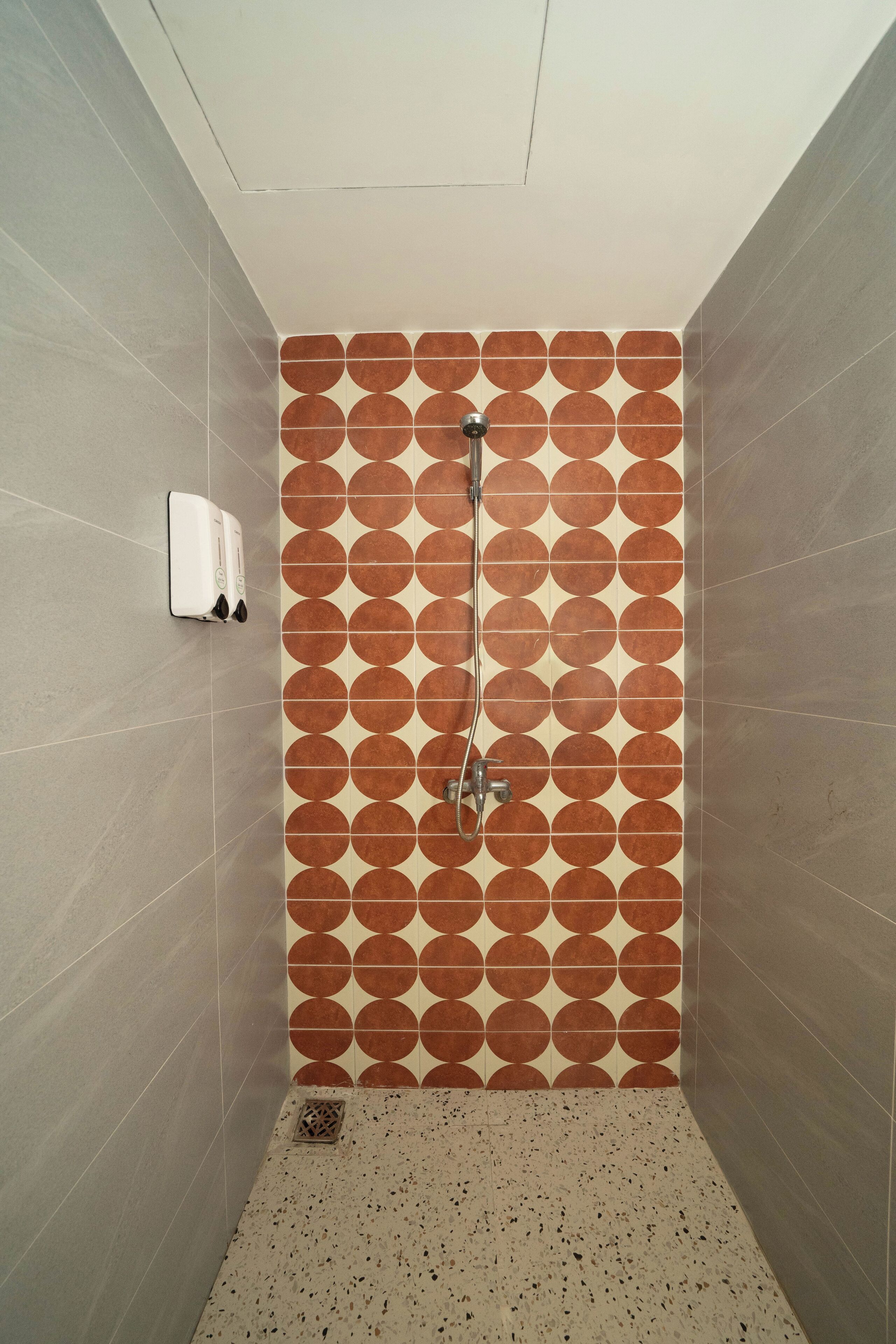 Une petite douche carrelée avec un motif géométrique sur le mur, une douchette à main et un distributeur de savon fixé au mur.