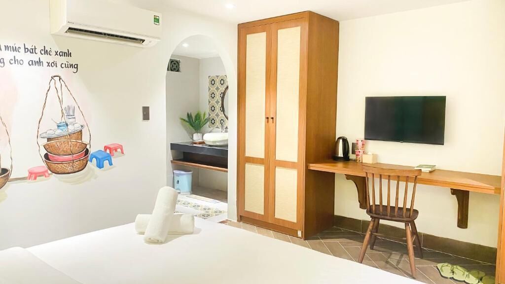 A bedroom with a bed, a wooden wardrobe, a desk with a chair, a television, and a wall-mounted air conditioner.